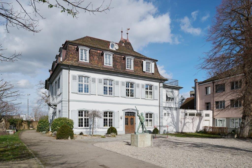 Large 2 story white house with brown roof, many windows, statue in front, walkway, small bushes