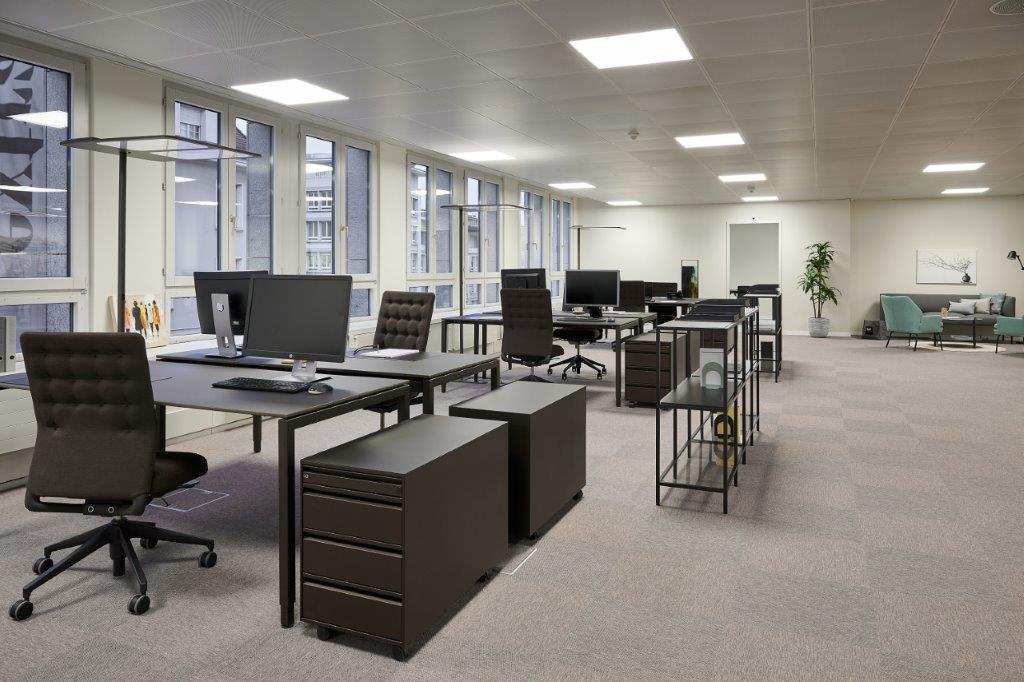 Large open office space with multiple workstations, desks, and computers. The room has large windows, bright lighting, and a modern, minimalist design. There are also some lounge chairs and a plant visible in the background.