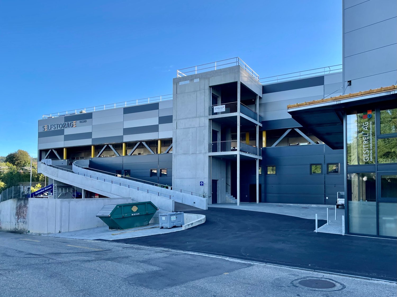 A large, modern commercial building with a gray and blue exterior. The building has multiple floors and a balcony on the upper level. There is a dumpster and parking space visible in the foreground.