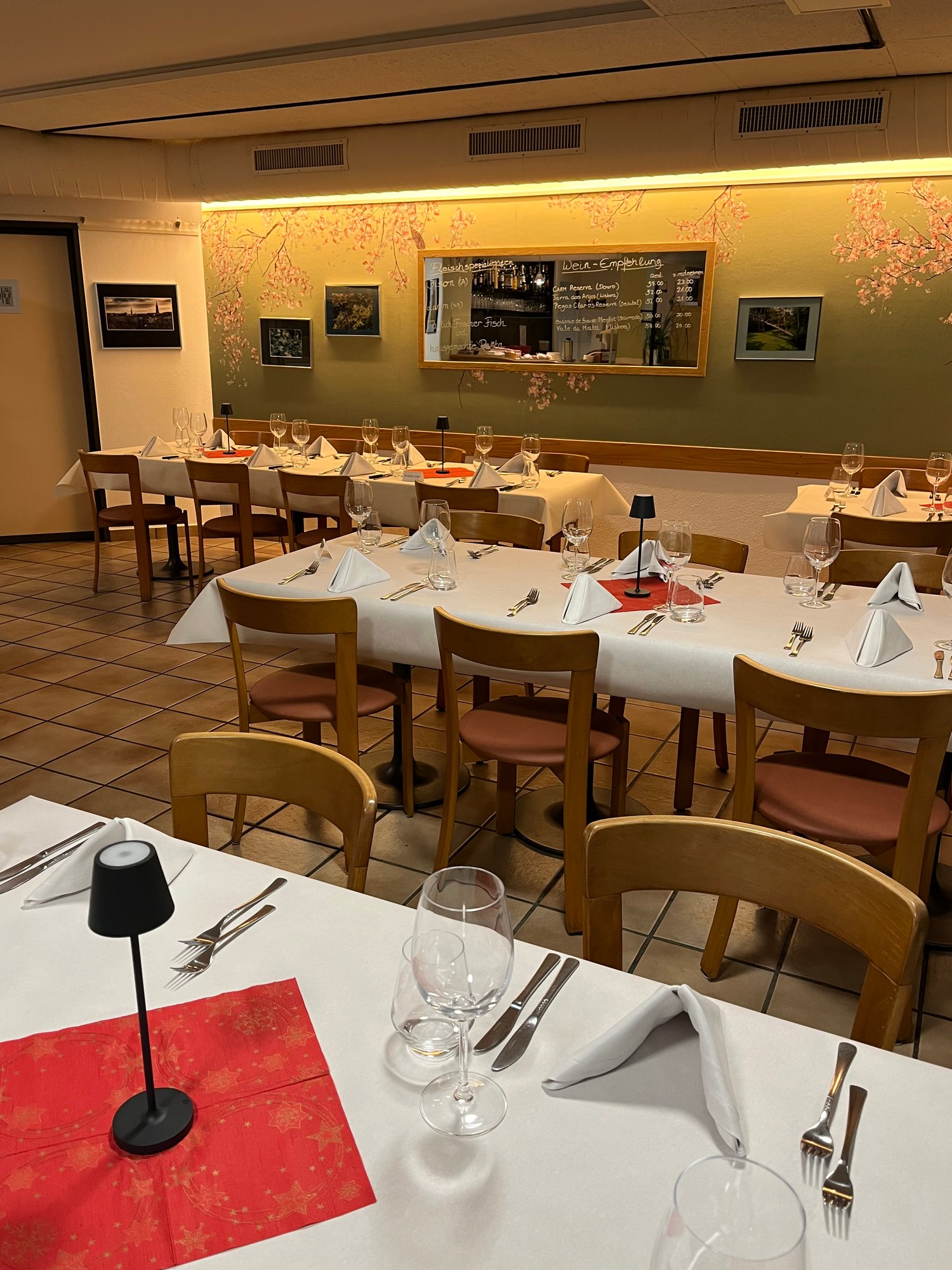 Set tables with white cloths and decorative napkins, several wooden chairs, menu board mounted on wall, lamp on table, patterned floor tiles