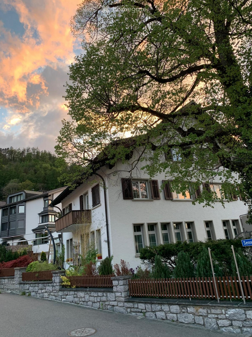 white facade house with brown wooden shutters, balcony on first floor, red roof, mountain in background