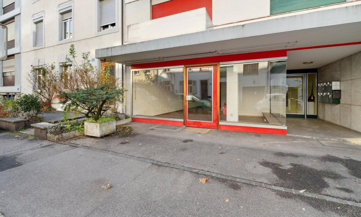 The entrance of an apartment building has large glass doors and windows. There are plants and greenery at the entrance and bicycles are parked on the side. A green car is parked in front.