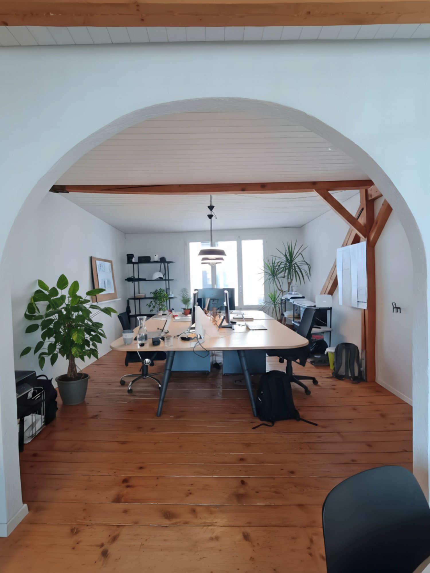 large room with wooden floor, two long tables, office chairs, desk, plant, desk lamp, framed picture, black bag, trolley, ceiling lamp