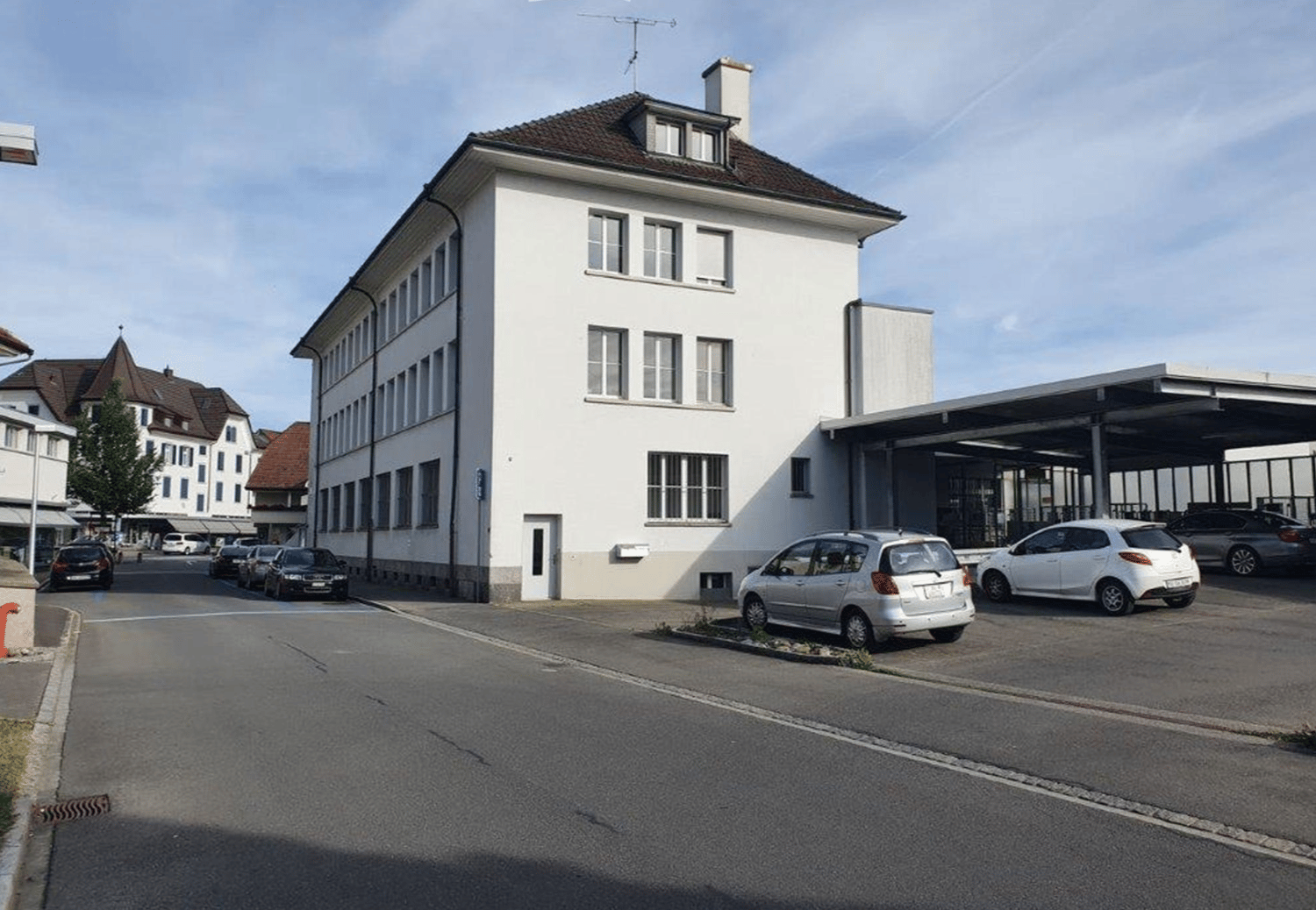 White building, two stories, multiple windows, parking in front, cars parked under carport.