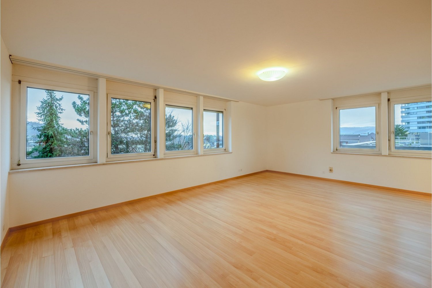 spacious, empty room with hardwood floor, white walls, three large windows with views, and a ceiling light