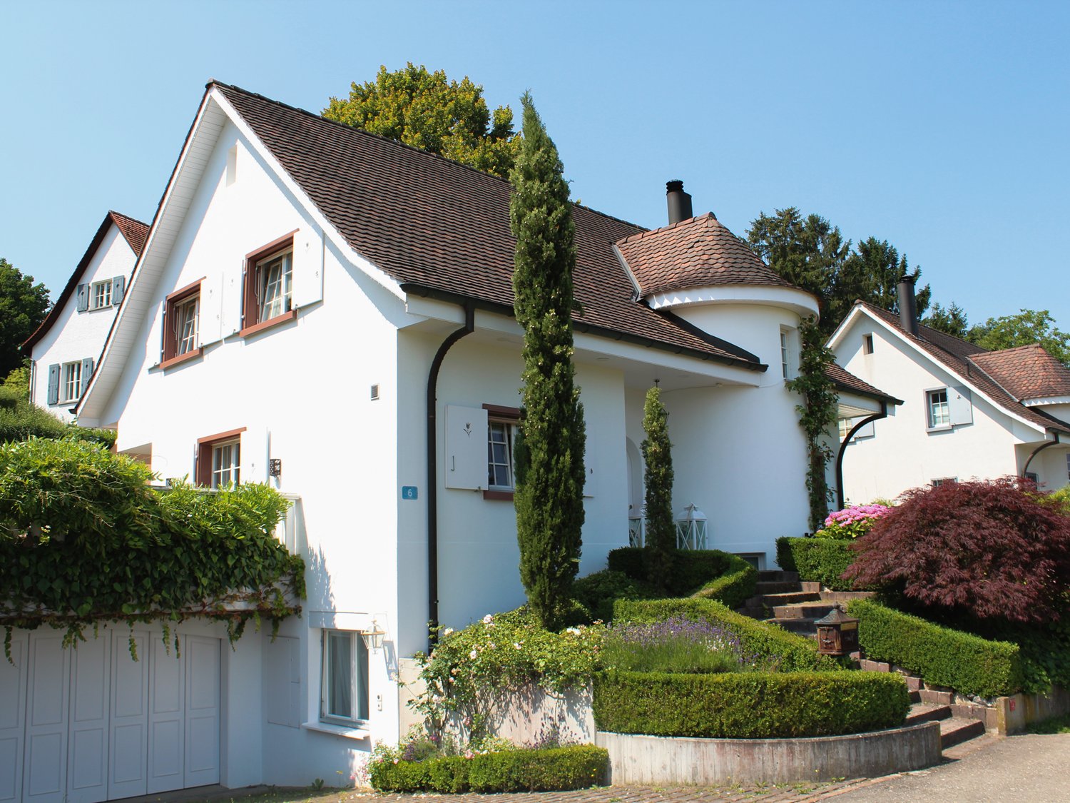 White house with brown roof, garage, brown trim, white shutters, front yard with well-kept shrubs, pathway, greenery