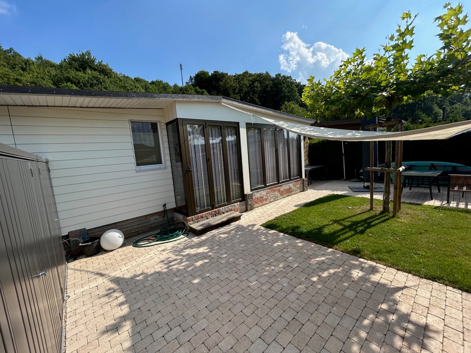 Single story house, white exterior, brick wall on the sides, green brick pathway, grassy lawn, patio with an awning and a green hose