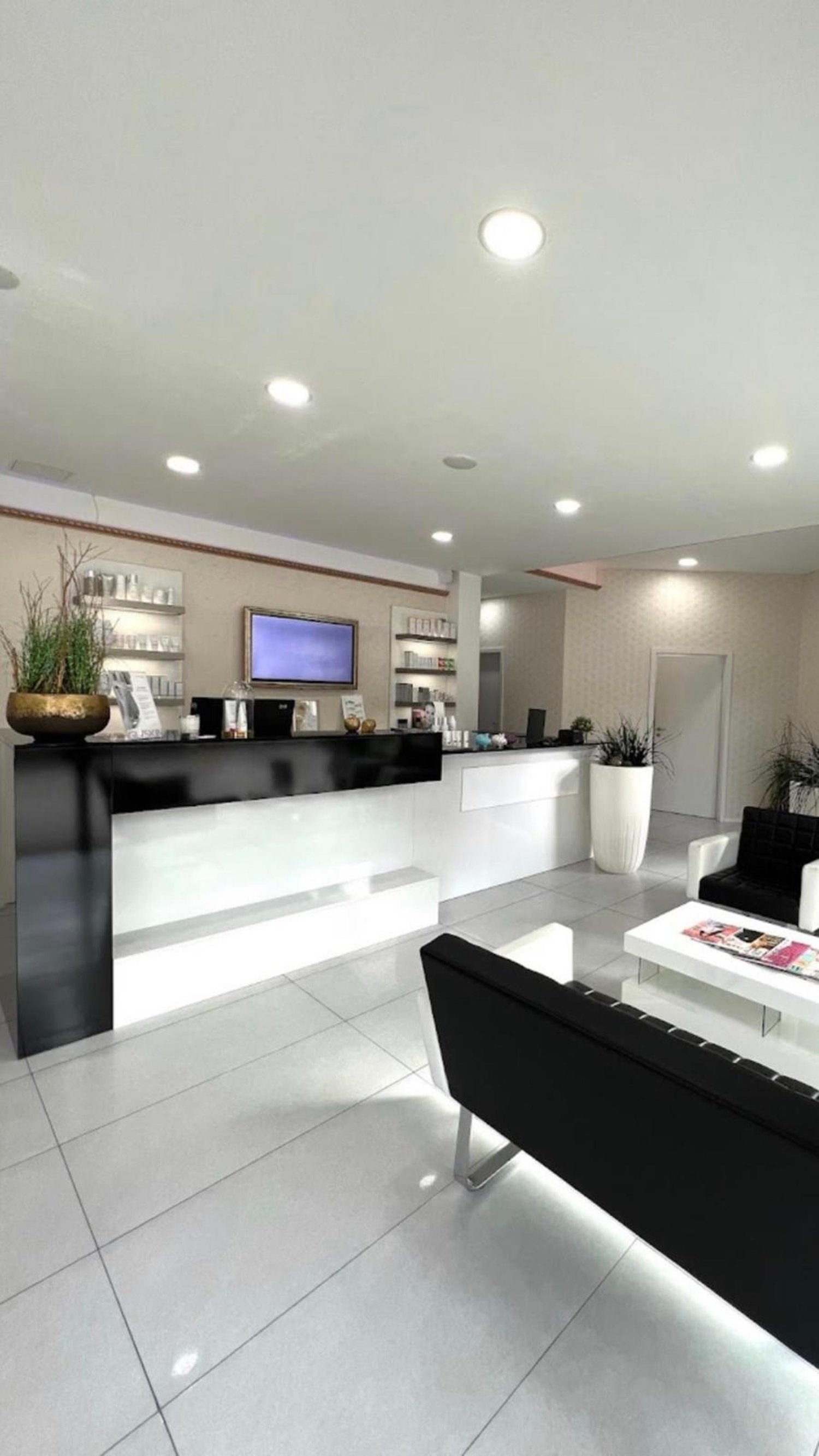 modern office reception area, white and black decor, reception desk, tv mounted on wall, shelves with products, two black sofas, white coffee table, plants