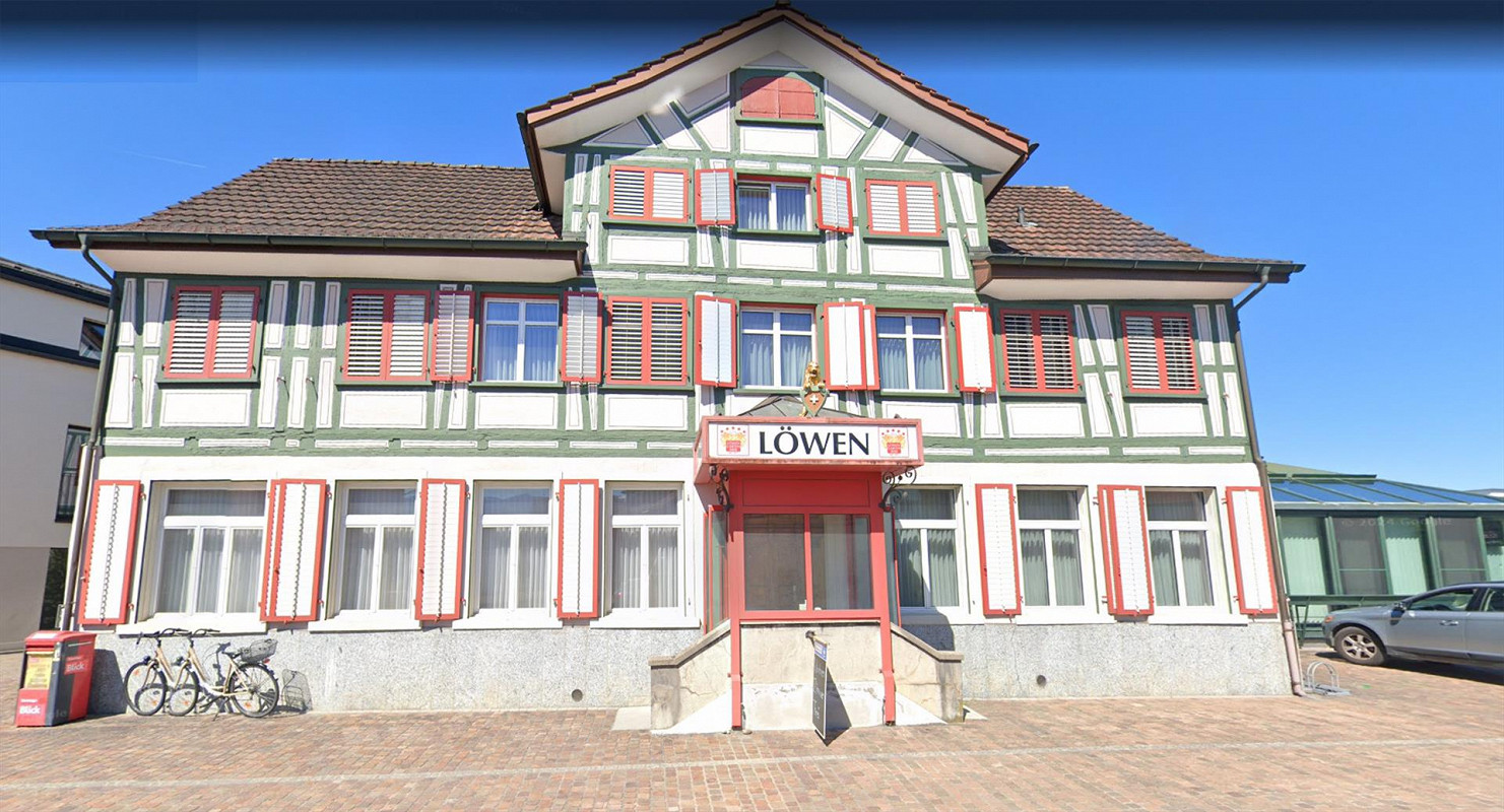 House: two story, 2 wings, brown roof, many windows, Löwen sign, red shutters