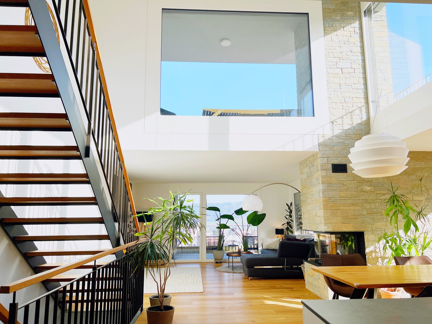 open floor plan, large windows, living room with couch, dining table, fireplace, staircase, wooden floor, plants