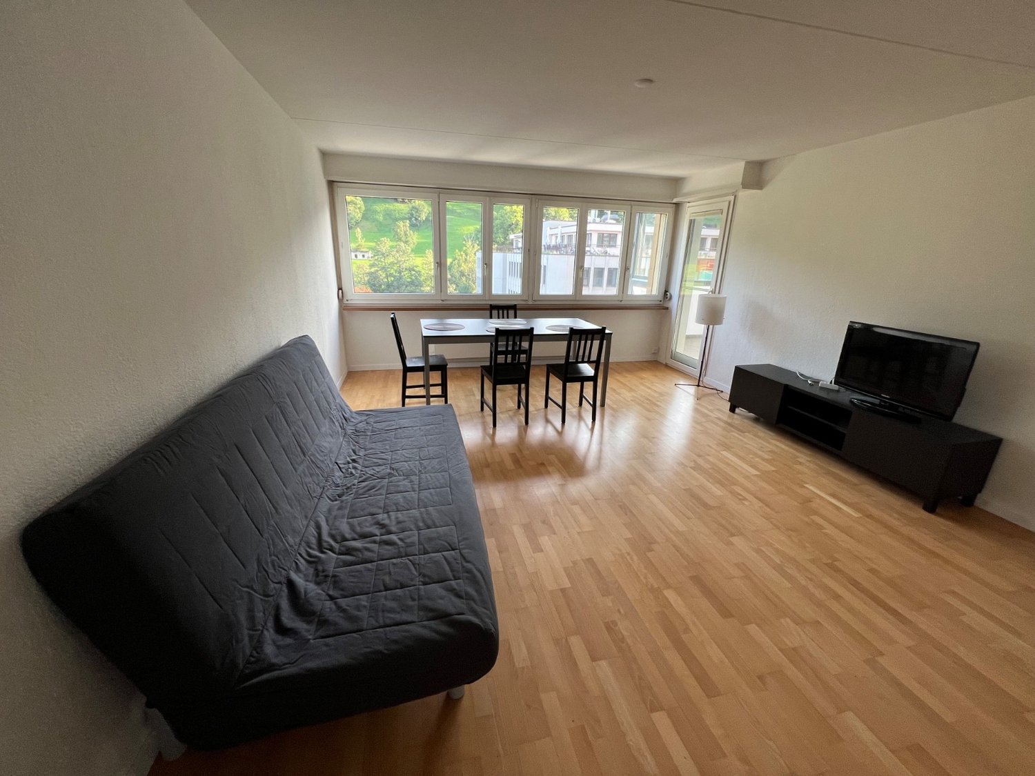 Spacious living room with wooden floors, modern furniture, dining table and chairs, black sofa, black television stand, large windows, white walls
