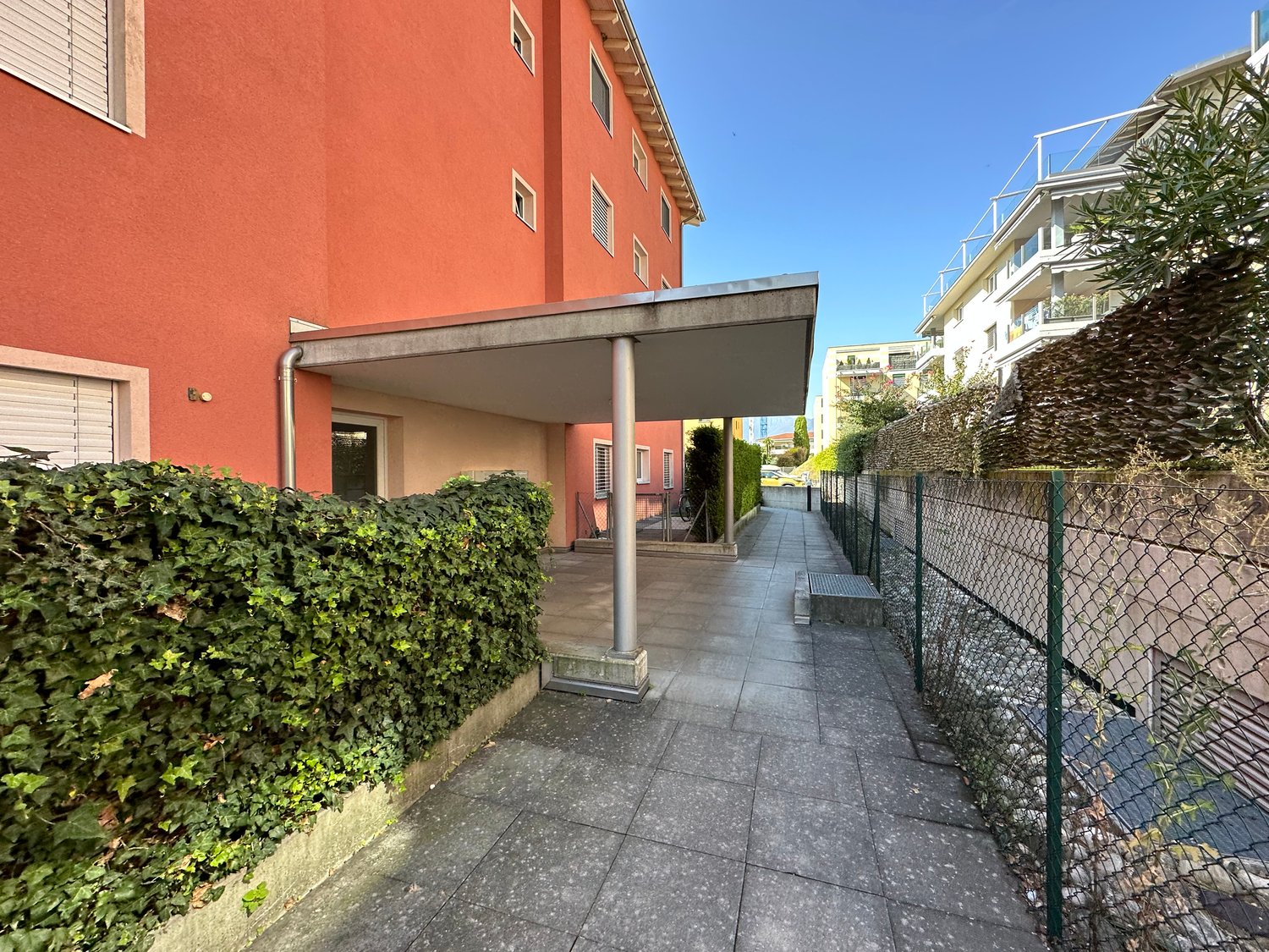 A covered parking area at the front of a building, tiled ground, fenced boundary with greenery, metal railings, multiple windows on building, side entrance