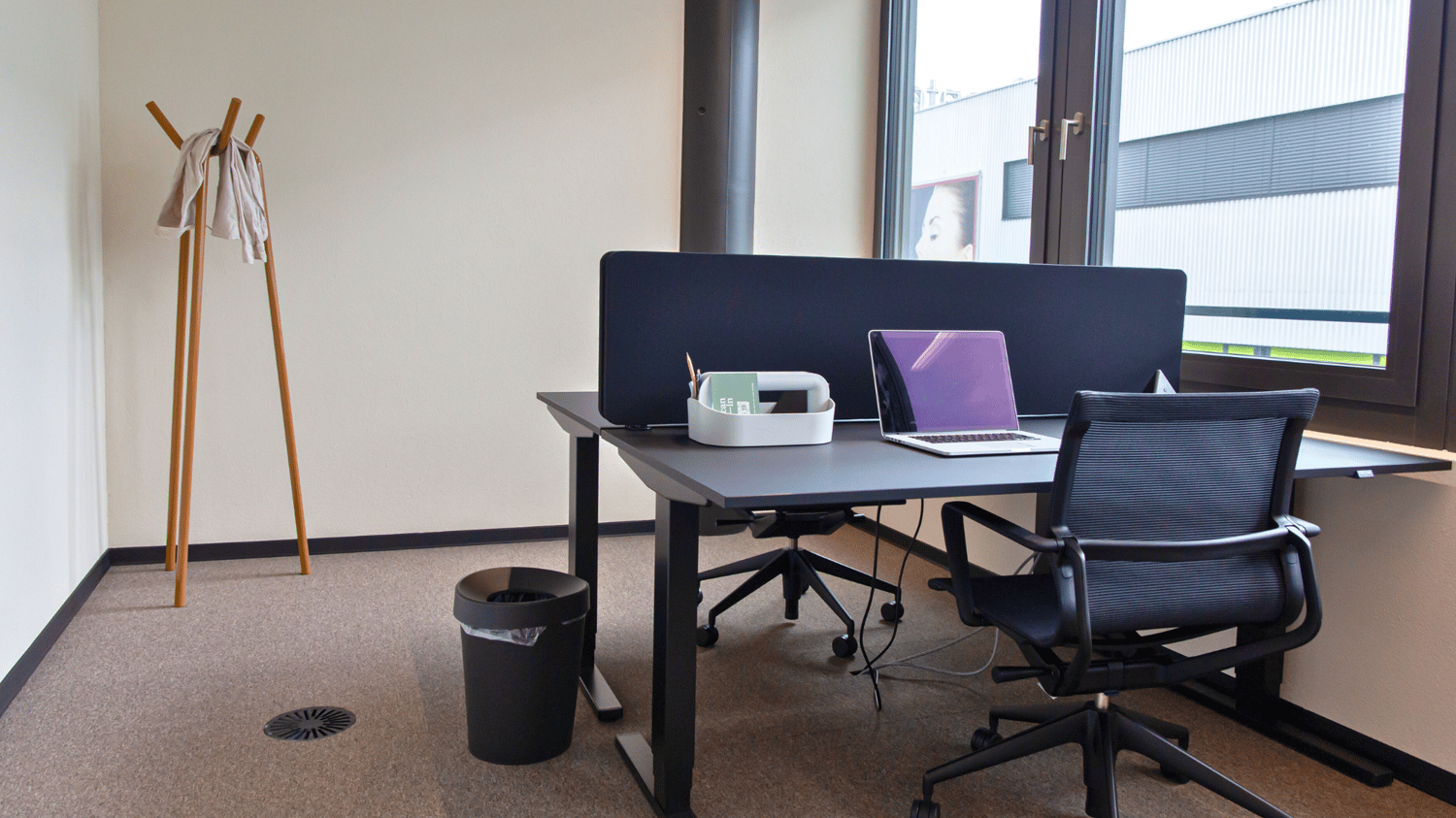 Office desk, office chair, laptop, trash bin, coat rack, large window with view outside