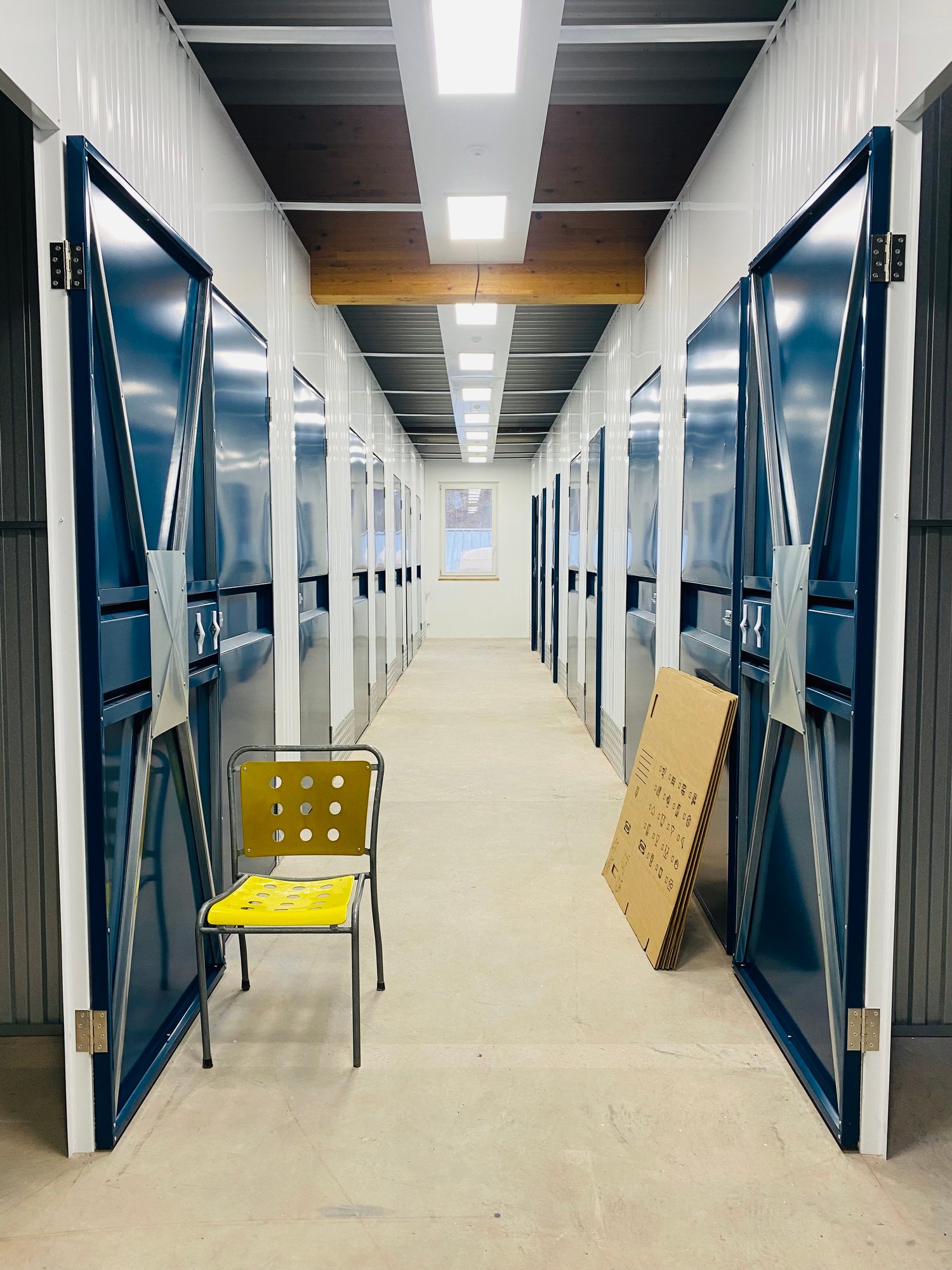 The image shows a long hallway with multiple storage units on both sides. The units have metal doors, and there is a yellow chair and a cardboard box in the hallway. The ceiling has lighting fixtures and the floor is a concrete surface.