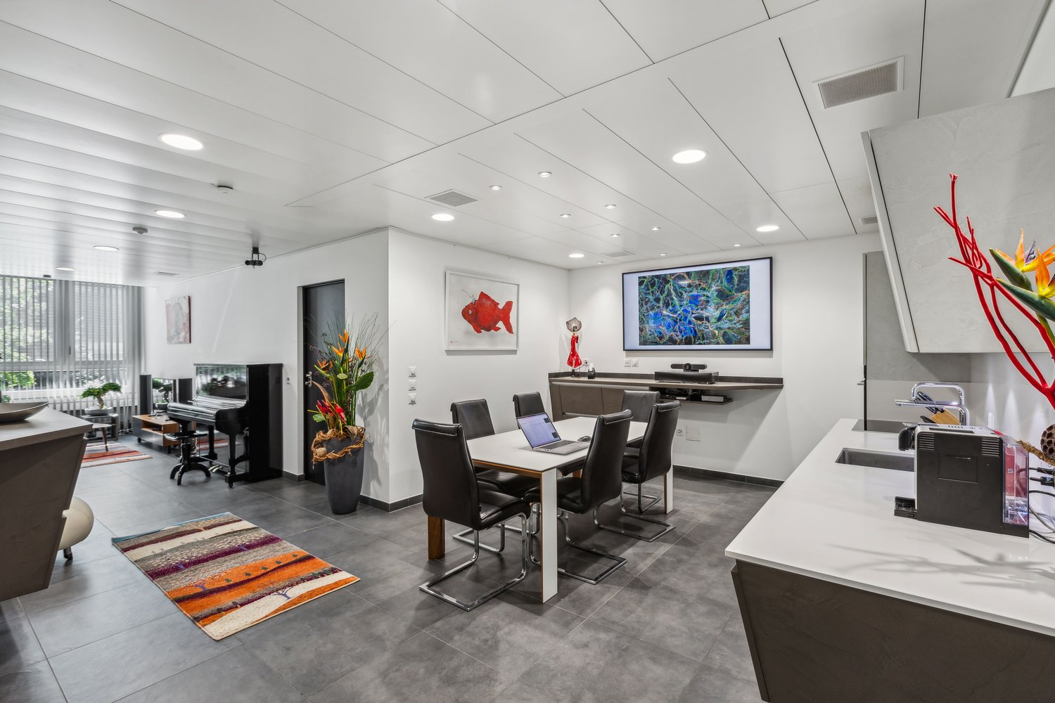 Open-concept living and dining area with modern, minimalist decor. The space features a large TV and entertainment system, a dining table with chairs, and a kitchen area with a dishwasher. The walls are decorated with colorful artwork, and the room has a